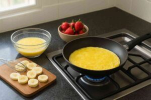 ಗೃಹ ಅಡುಗೆಮನೆಯಲ್ಲಿ ಗ್ಯಾಸ್ ಸ್ಟೌವ್ ಮೇಲೆ ಬೇಯುತ್ತಿರುವ ಬಾಳೆಹಣ್ಣು ಓಮ್ಲೆಟ್, ಪಕ್ಕದಲ್ಲಿ ಬಾಳೆಹಣ್ಣಿನ ತುಂಡುಗಳು, ಸ್ಟ್ರಾಬೆರಿ ಮತ್ತು ಮೊಟ್ಟೆಯ ಮಿಶ್ರಣದ ಬಟ್ಟಲು ಇರಿಸಿರುವ